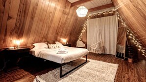 A cozy attic bedroom with wooden walls, a bed with white linens, a hanging chair, and string lights.