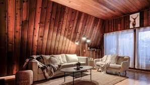 A cozy living room with wooden paneling, a sofa, two armchairs, a coffee table, and a large window with curtains.