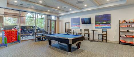 A pool table in a well-lit room with a carpeted floor, chairs, and a bookshelf with magazines.