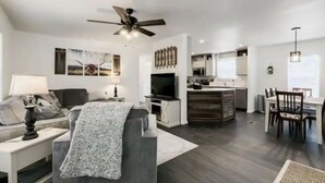 A modern living room with a grey sofa, a coffee table, and a TV. There is a ceiling fan, a dining area with a table and chairs, and a kitchen with cabinets and a countertop. - Cozy Mountain Town Farmhouse for Winter Fun in Flag!  (Flagstaff)