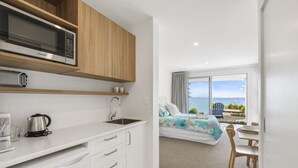 Cocina moderna con microondas, hervidor y lavabo. Se ve un dormitorio con cama y vista al mar a través de una puerta abierta.