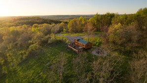 A cabin with a deck, surrounded by forested hills and a clear sky.