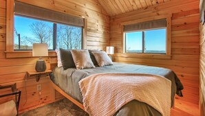 A wooden cabin bedroom with a bed, two windows, a lamp, and a nightstand.