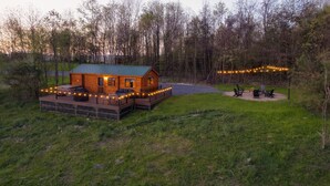 A wooden cabin with a deck, surrounded by trees and a grassy area with outdoor seating. - Sanctuary Summit | Hot Tub, Panoramic Views, ADA Friendly Cabin (Brookville)