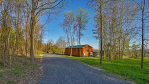 A gravel road leading to a wooden cabin with a green roof, surrounded by trees and grassy fields. - Sanctuary Summit | Hot Tub, Panoramic Views, ADA Friendly Cabin (Brookville)