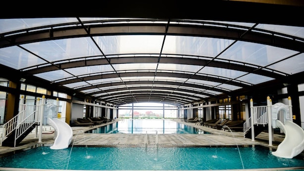 An indoor swimming pool with two slides, a glass ceiling, and a view of buildings outside.