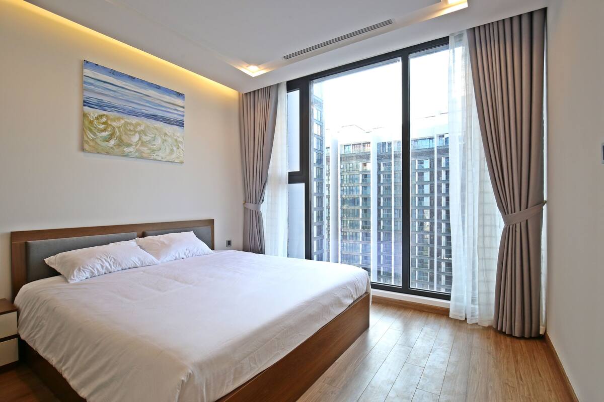 A modern hotel room with a large bed, a city view through floor-to-ceiling windows, and a painting on the wall.