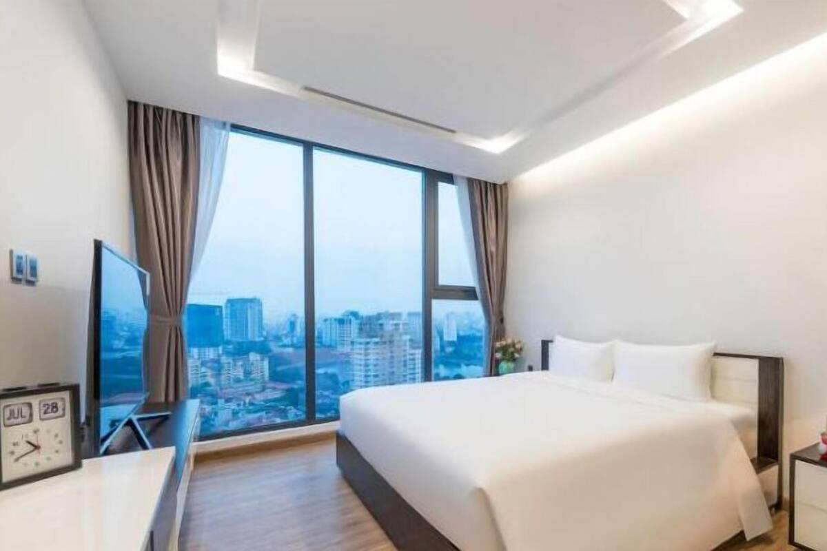 A modern hotel room with a large bed, a bedside table with a lamp, a TV, and a view of the cityscape through large windows.