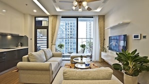 A modern living room with a sofa, coffee table, and TV. There is a kitchen area with a kettle and cabinets. Large windows provide a view of high-rise buildings.