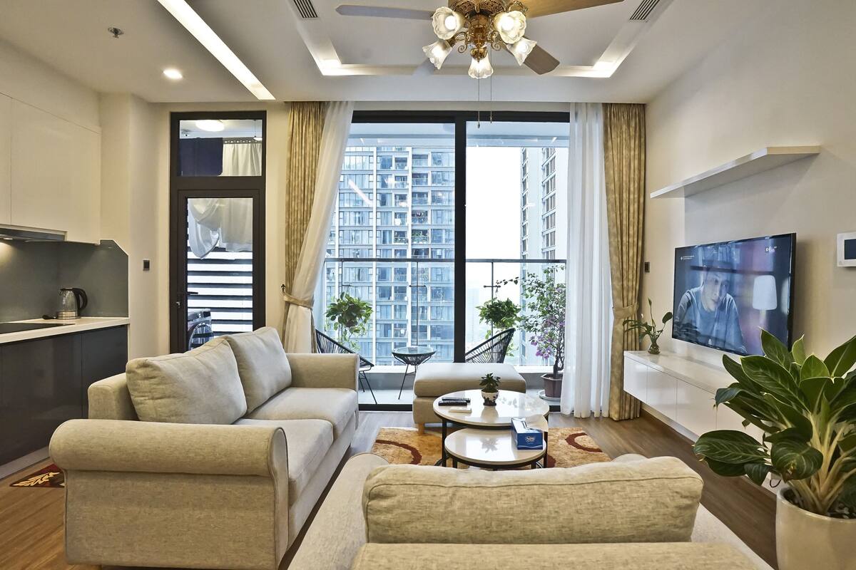 A modern living room with a sofa, coffee table, and TV. There is a kitchen area with a kettle and cabinets. Large windows provide a view of high-rise buildings.