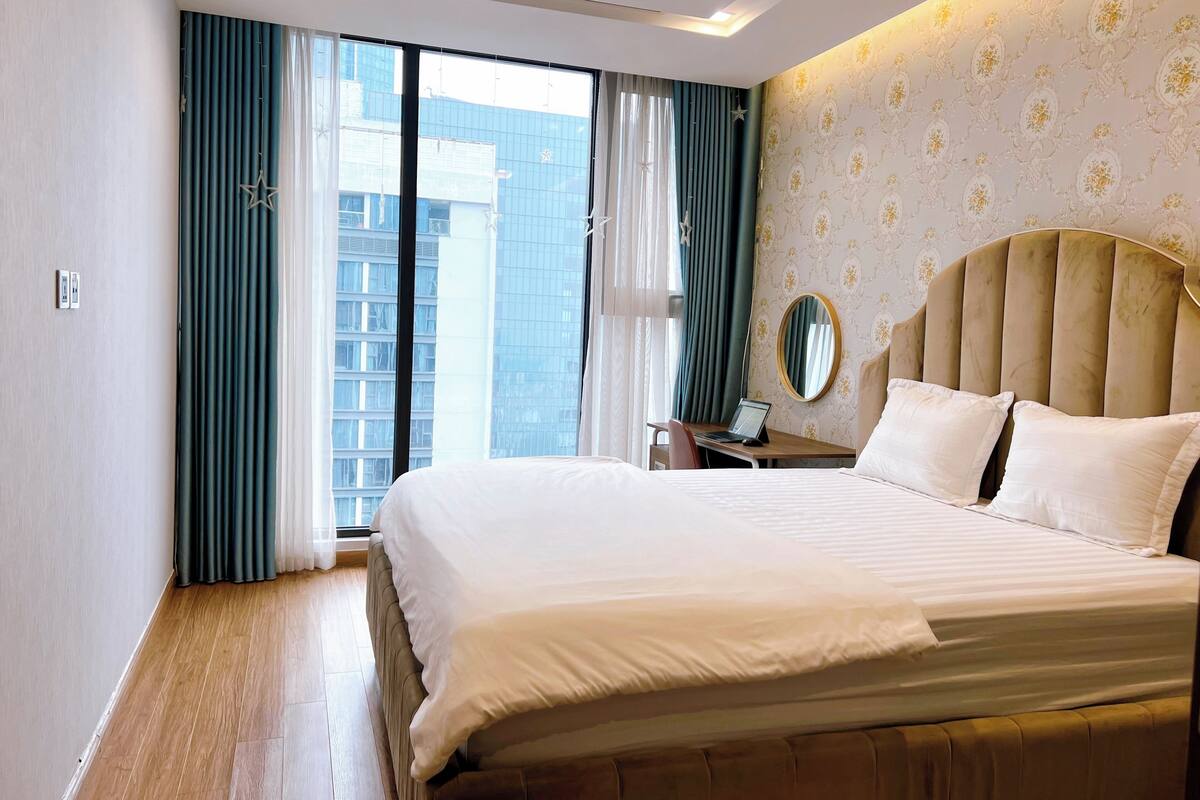 A hotel room with a large bed, a desk with a laptop, a mirror, and a view of a cityscape through floor-to-ceiling windows.