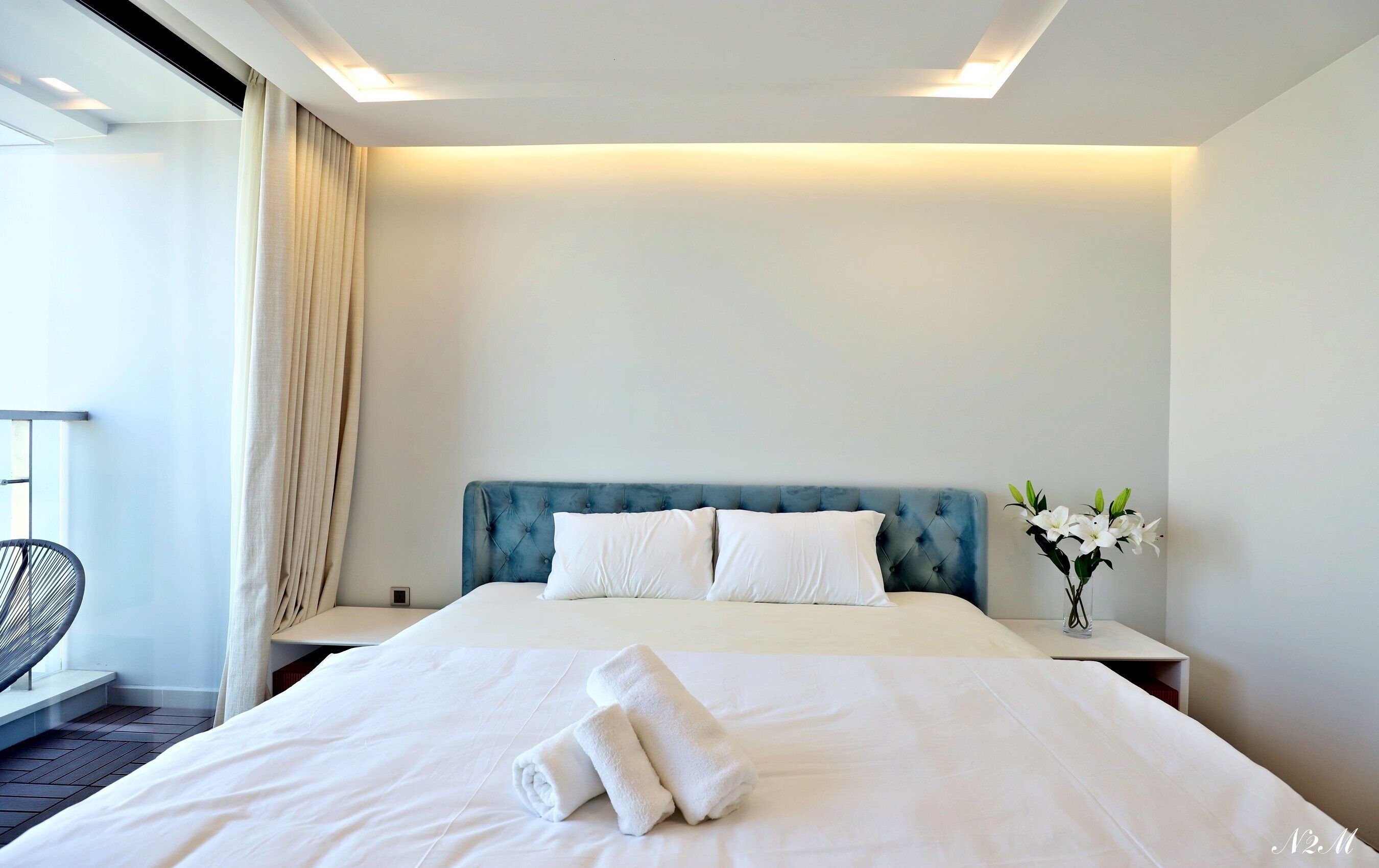 A hotel room with a large bed, two pillows, a nightstand with a vase of flowers, and a chair by the window.