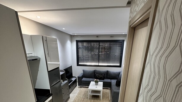 A modern kitchen with a built-in oven and microwave, a grey sofa with a coffee table, and a large window with blinds. - Ziad Studio Moderne (Casablanca)
