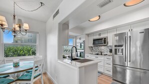Una cocina moderna con armarios blancos, electrodomésticos de acero inoxidable, una isla central con pileta y un comedor con sillas color turquesa.