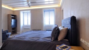 Une chambre avec un grand lit, un ventilateur de plafond, une bibliothèque et deux fenêtres.