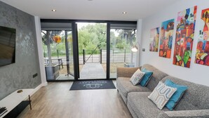 A modern living room with a sofa, a coffee table, and a TV mounted on the wall. There is a glass door leading to an outdoor area with a bench and a lamp. - The Tunnel at Bridge Lake Farm and Fishery (Banbury)