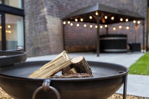 A fire pit with logs, a brick building, and string lights in the background. - The Tunnel at Bridge Lake Farm and Fishery (Banbury)