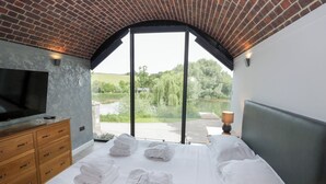 A bedroom with a large bed, a TV, wooden drawers, and a view of a lake through large windows. - The Tunnel at Bridge Lake Farm and Fishery (Banbury)