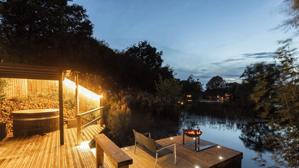 A wooden deck with a hot tub, seating, and a fire pit by a calm lake at dusk. - The Tunnel at Bridge Lake Farm and Fishery (Banbury)