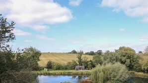 A serene pond with a wooden dock, surrounded by lush greenery and a small cabin in the distance.