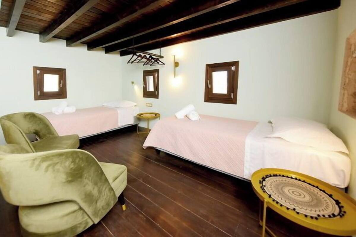 Quarto com duas camas, uma cadeira, uma mesinha e um cabideiro.