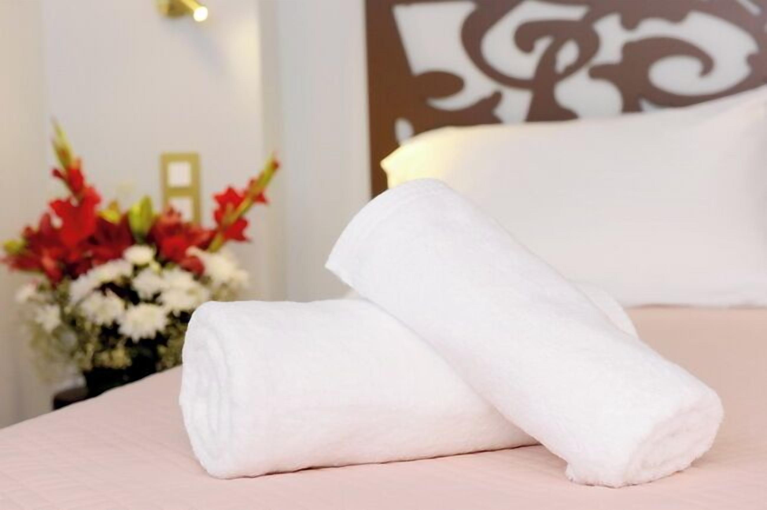 Una cama impecable con toallas blancas y un jarrón con flores rojas y blancas al fondo.