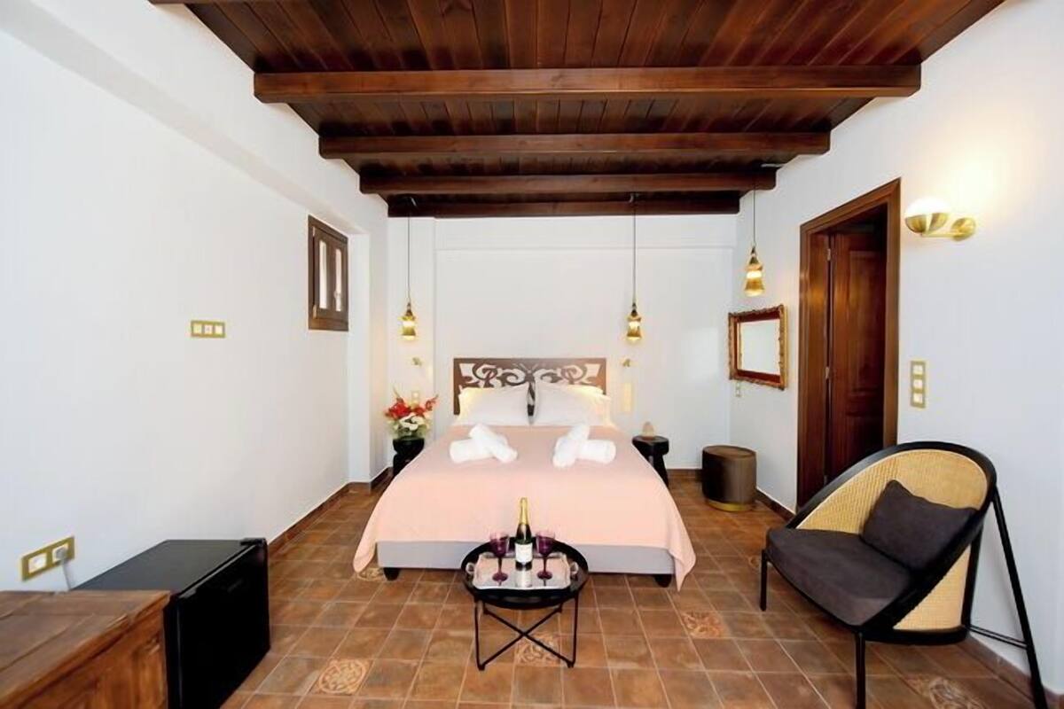 Quarto com cama, mesas de cabeceira, uma cadeira, uma pequena mesa com garrafa e copos, e teto de madeira.