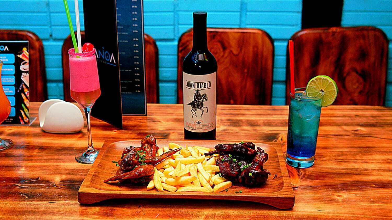 A wooden table with a plate of food, a bottle of wine, and two drinks.