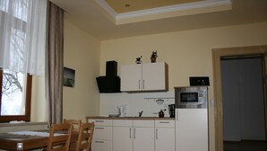 Cocina pequeña con armarios blancos, microondas y hervidor. Se observa una mesa de comedor con sillas y una ventana con cortinas.