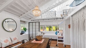 A spacious room with a wooden dining table, a chandelier, a bed with a white canopy, and a view of greenery outside.