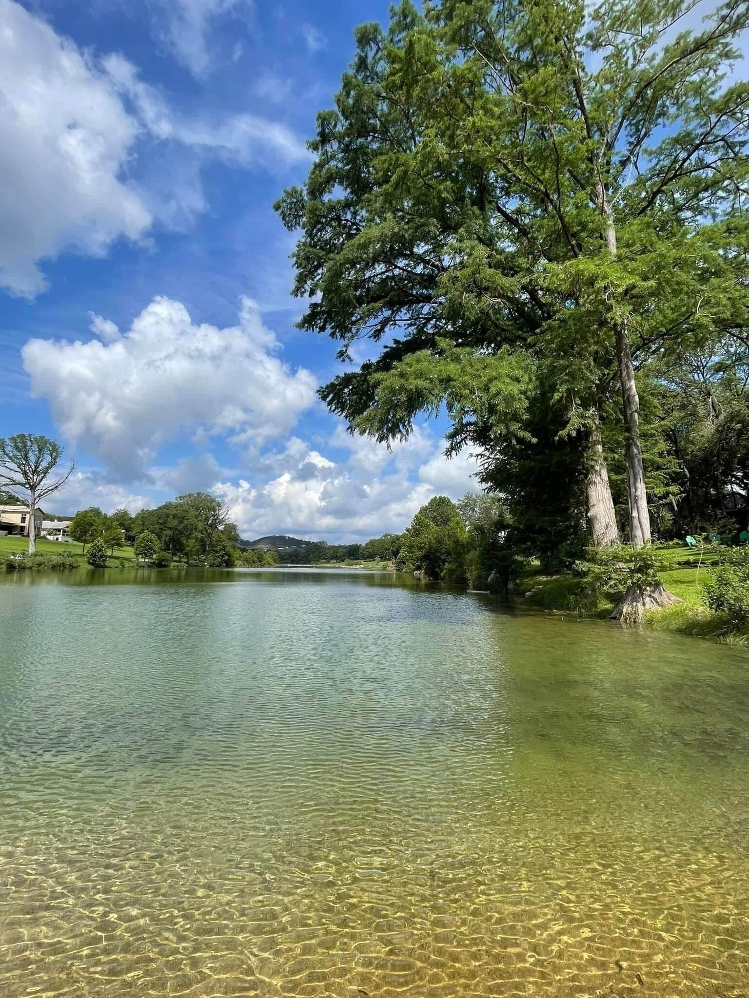 A clear lake surrounded by trees and a building in the distance.