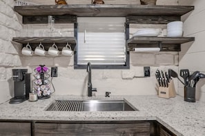 A kitchen with a stainless steel sink, wooden shelves, a coffee maker, and a knife block. - The Mockingbird Inn | Private Hill Country Cabin (Wimberley)