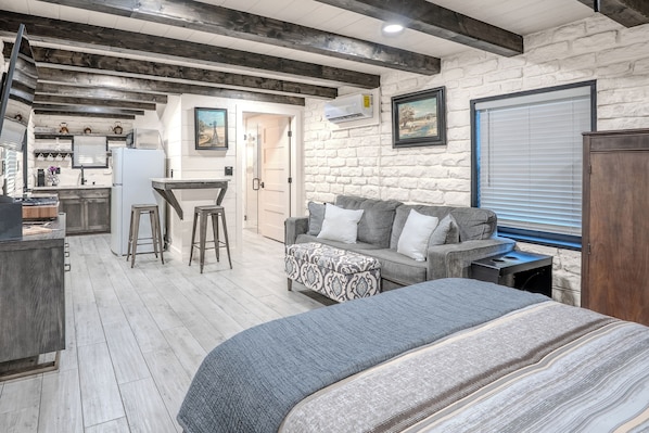 A room with a bed, a sofa, a kitchen area, and a stone wall. - The Mockingbird Cabin - Peaceful Hill Country Getaway (Wimberley)