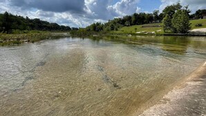 A clear river with a forested hillside and a building in the background.