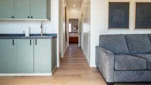 A modern kitchen with light green cabinets, a dark countertop, and a wooden floor. A grey sofa is positioned in the foreground, and a hallway leads to a bedroom with a bed and a painting on the wall. - Courtyard Casita - 2bed/2bath Inn (Torrey)