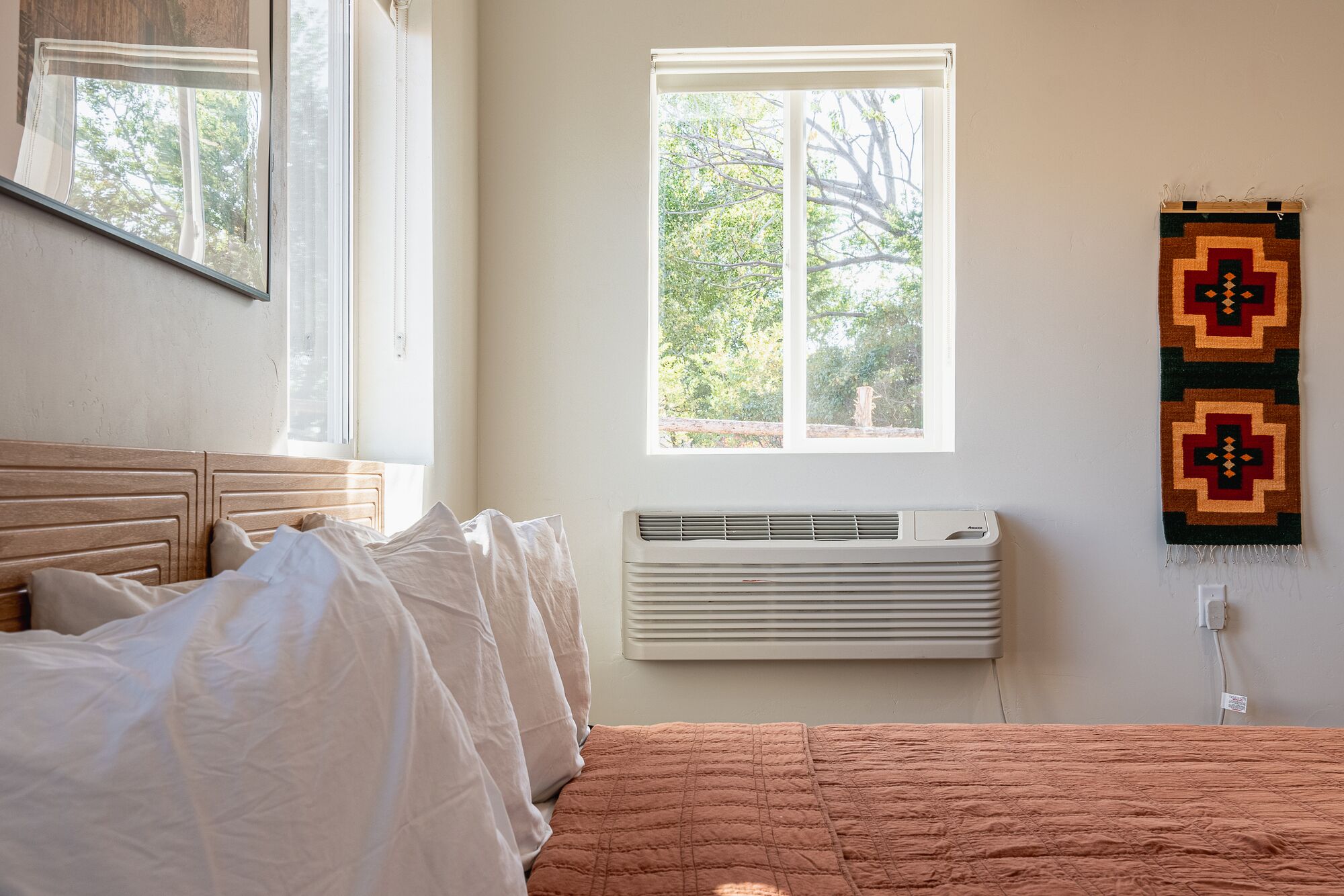 A bedroom with a bed, a window, a wall-mounted air conditioner, and a wall hanging.