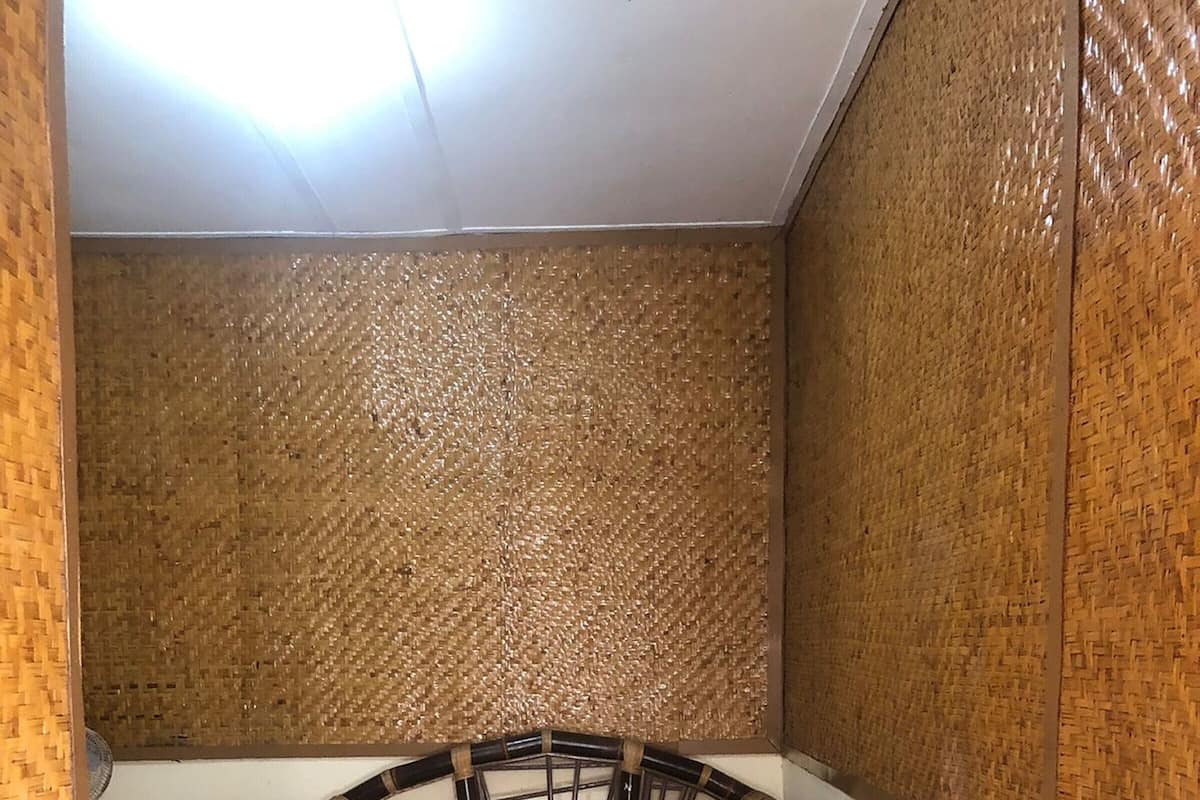 A small, narrow room with a single bed, a wooden headboard, and a woven wall panel.