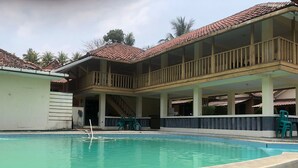 Un área de piscina con cubierta de madera, un espacio de estar techado con barandilla y un edificio con tejado de azulejos.