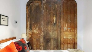 Een kamer met een houten garderobe, een lijst met een bord op de muur, een bed met een witte dekbedovertrek en een rood en zwart kussen.