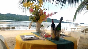 Une table dressée pour deux personnes, avec une nappe jaune, un vase de fleurs et deux bouteilles de champagne, sur une plage offrant une vue sur la mer et les montagnes.