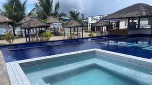 Une piscine dotée d’une terrasse carrelée, entourée de palmiers et bordée de constructions à toits de chaume.