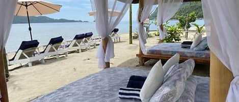 A beachside bed with a patterned bedspread and pillows, a canopy overhead, and beach chairs and umbrellas in the background.