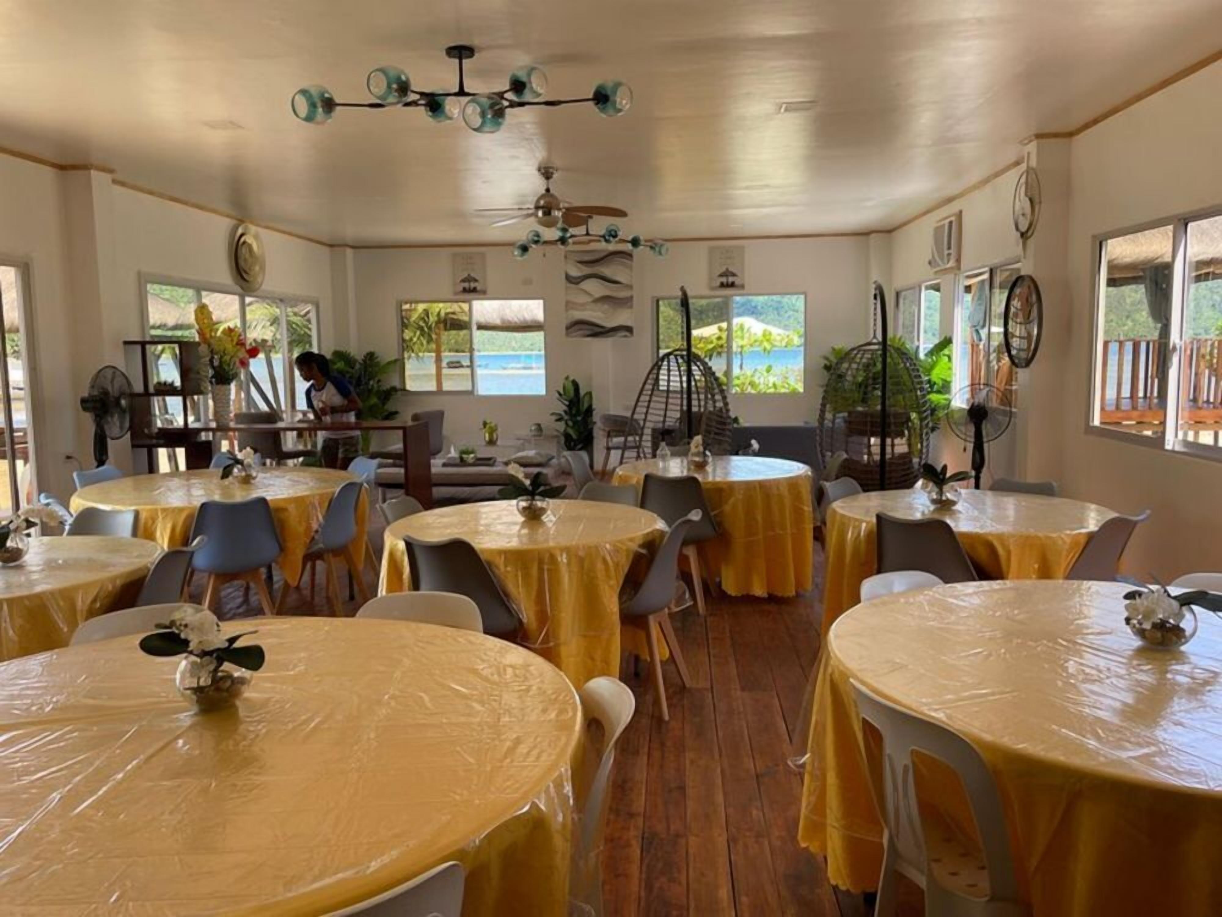 Un ristorante con tavoli rotondi ricoperti da tovaglie gialle, una ventola a soffitto e vista sull'esterno.