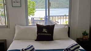 Una cama con una colcha azul, dos toallas dobladas y un cojín con el texto 'BURBERRY'. Una ventana muestra una vista de un lago y colinas.