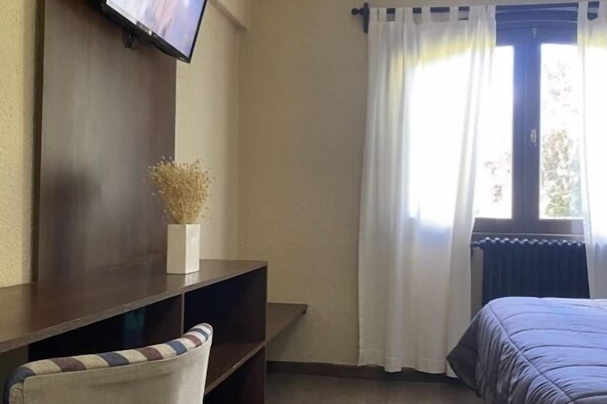 A hotel room with a bed, a chair, a TV mounted on the wall, and a window with curtains.