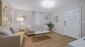 A living room with a beige sofa, a coffee table, a floor lamp, and a door with a 'No Smoking' sign. - Private Apartment on River Ave. close to main st (Patchogue)
