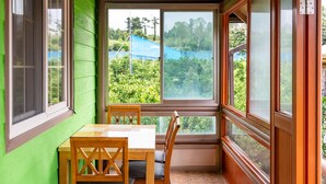 A corner room with a table and chairs, large windows, and a view of greenery outside. - Jeju Jeonwonsaenghwal Diorem Pension (Jeju City)