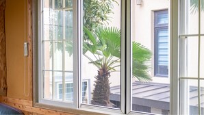 A window with a view of a palm tree and a building with a blue window frame. - Jeju Jeonwonsaenghwal Diorem Pension (Jeju City)