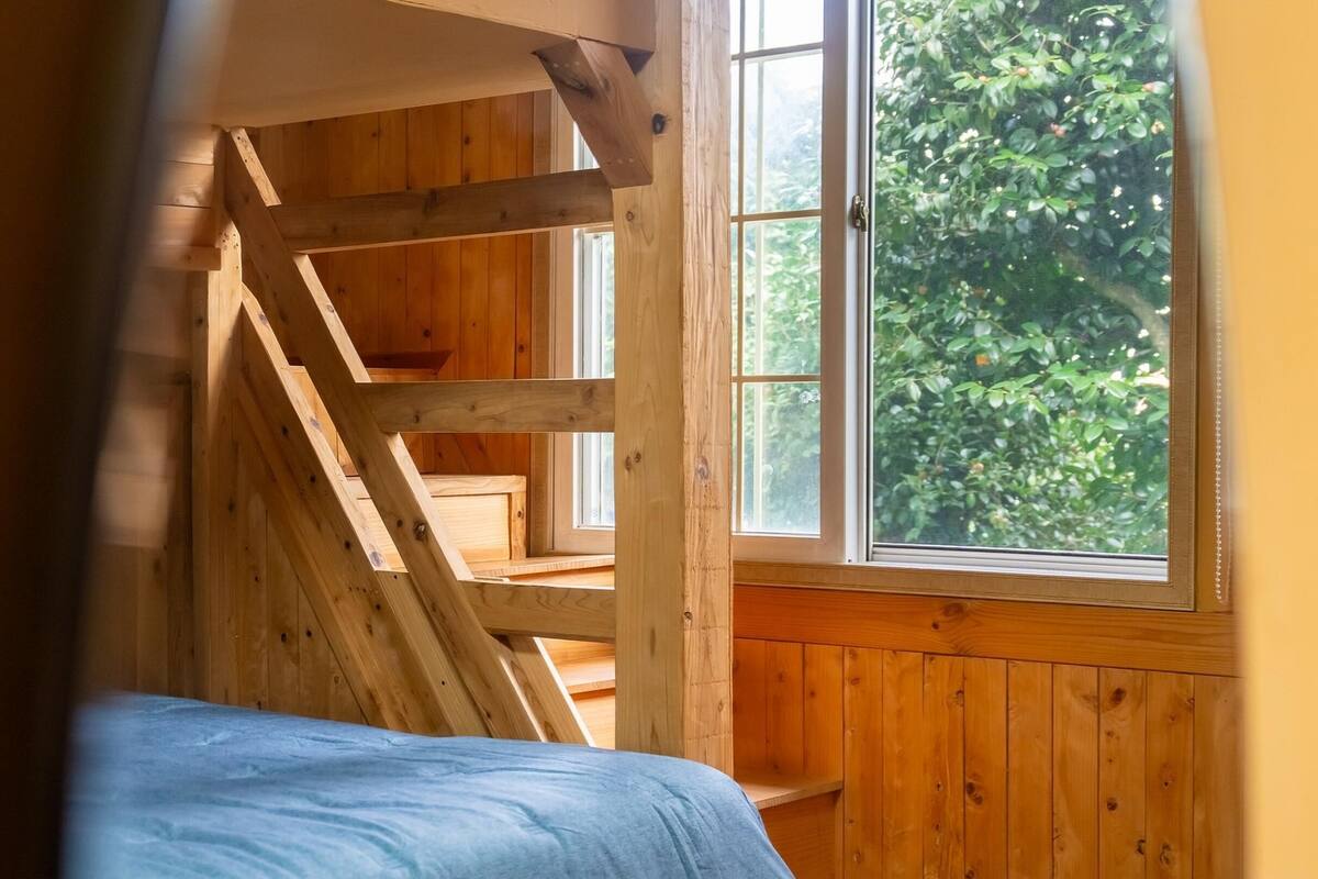 Una habitación con techo de madera inclinado, una ventana con vistas a la vegetación y una cama con cubrecama azul.