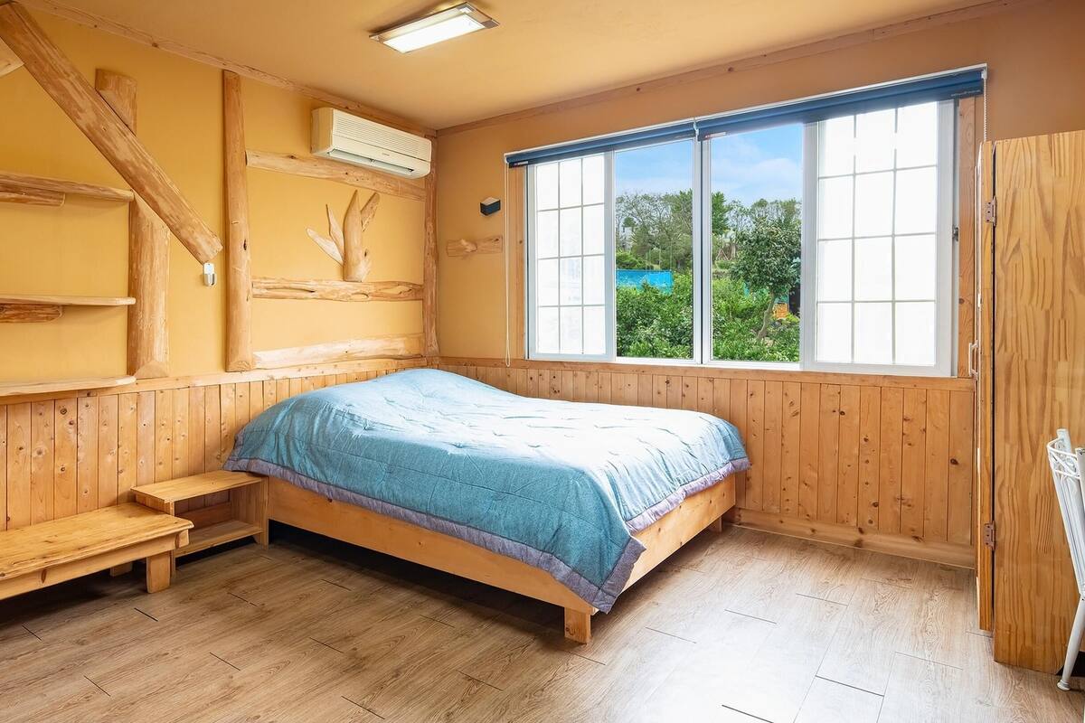 Un dormitorio con una cama de madera, un banco, una ventana con vistas a la vegetación y un equipo de aire acondicionado.
